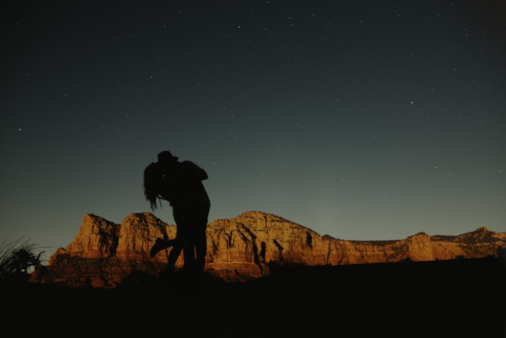A man holds a woman and tilts her back while kissing her with desert red rocks displayed in the background and crystal clear views of the stars in the sky.