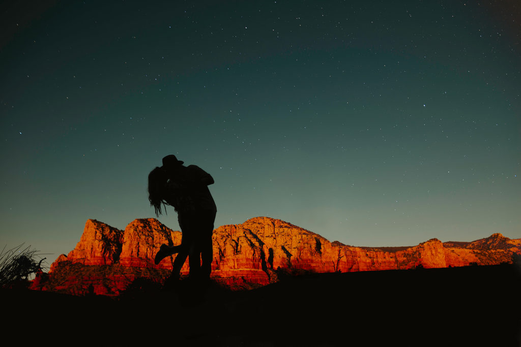 A couple embrace each other at the end of the day during their sunset elopement. The stars are beginning to appear in the sky as the sun casts a red and orange glow over the red rocks in the desert.