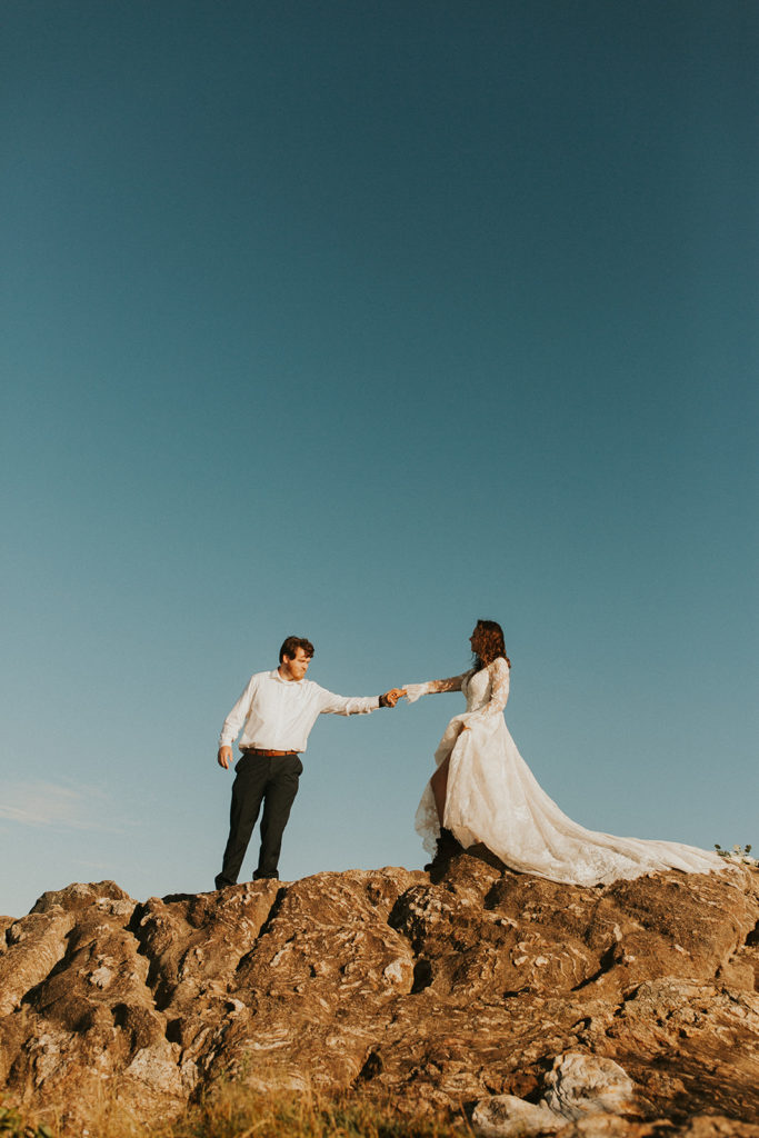 a husband and wife in wedding attire walk along a mountain ridge while holding out their hands to each other
