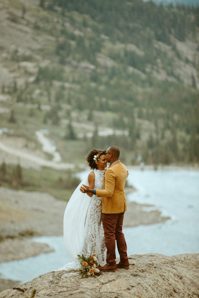 a couple in wedding attire dance along a cliff in front of an alpine lake in Colorado