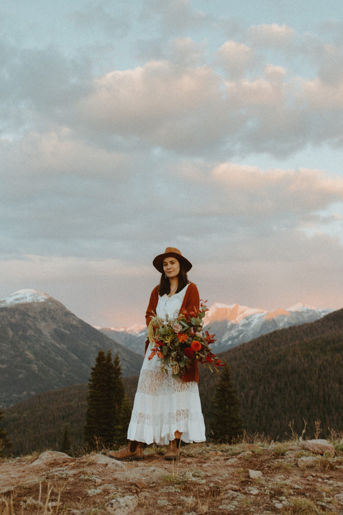 woman in a wedding dress holding a bouquet stands on top a mountain