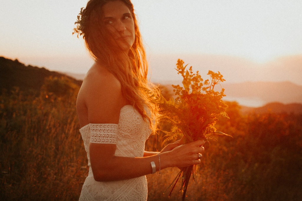 woman in a wedding dress holds a bouquet of flowers while smiling in front of a sunrise