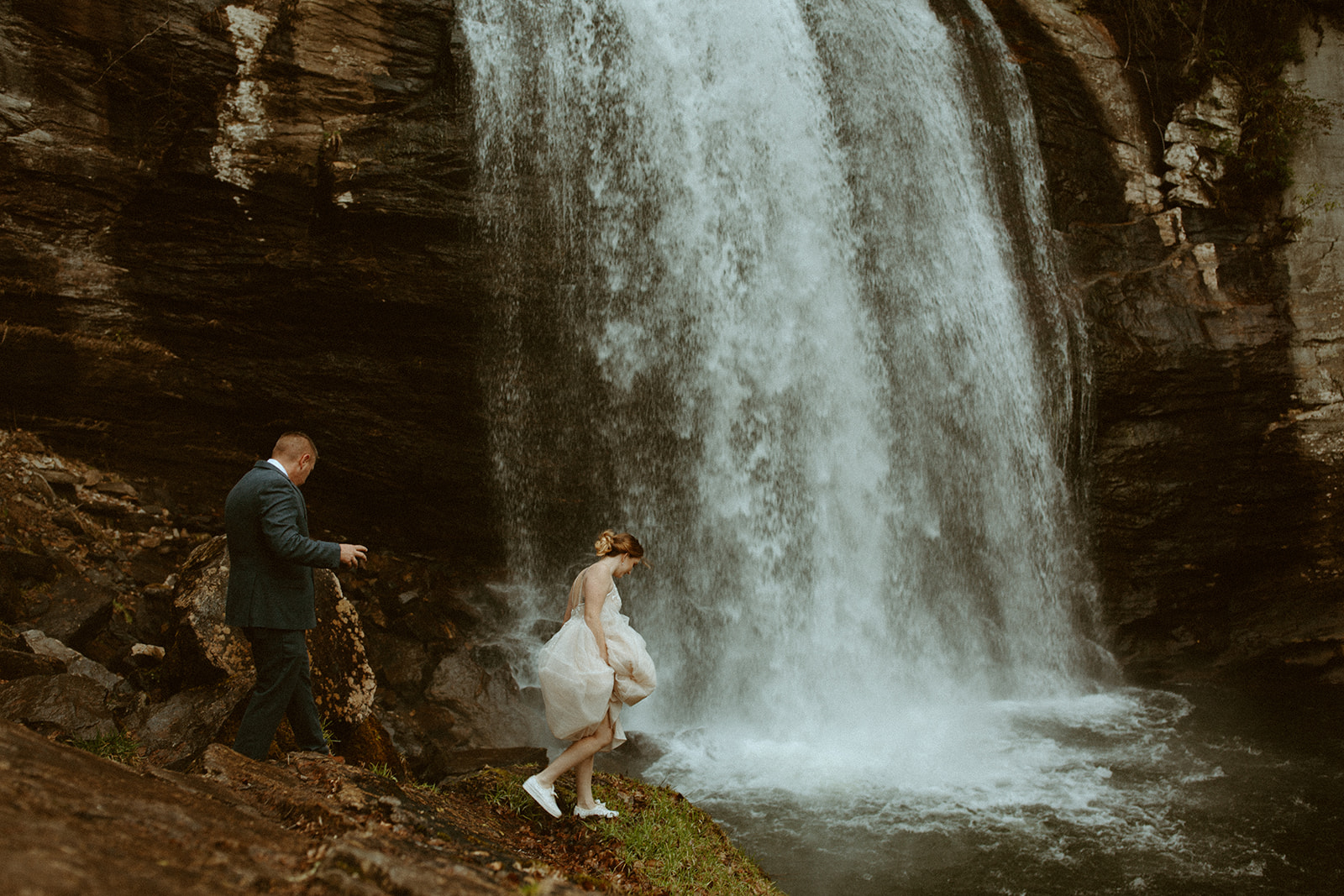 Husband and wife tread over rocks near a flowing waterfall in wedding attire