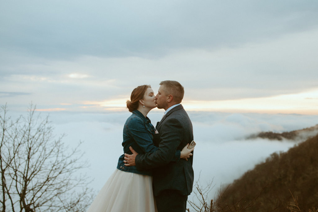 husband and wife kissing on top of a mountain during sunrise with cloud inversions in the background