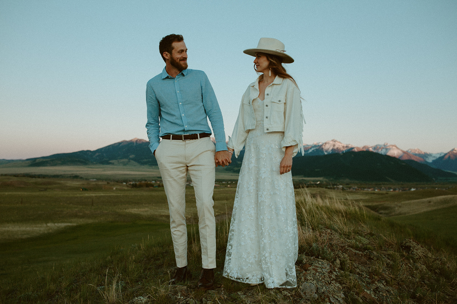 A man and woman in wedding attire stand in a field holding hands with mountains and a sunset in the background.