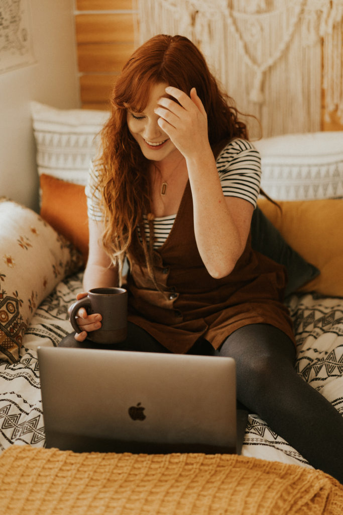 Girl sitting on a bed with a coffee mug in her hand looking down at her computer with her other hand combing through her hair.