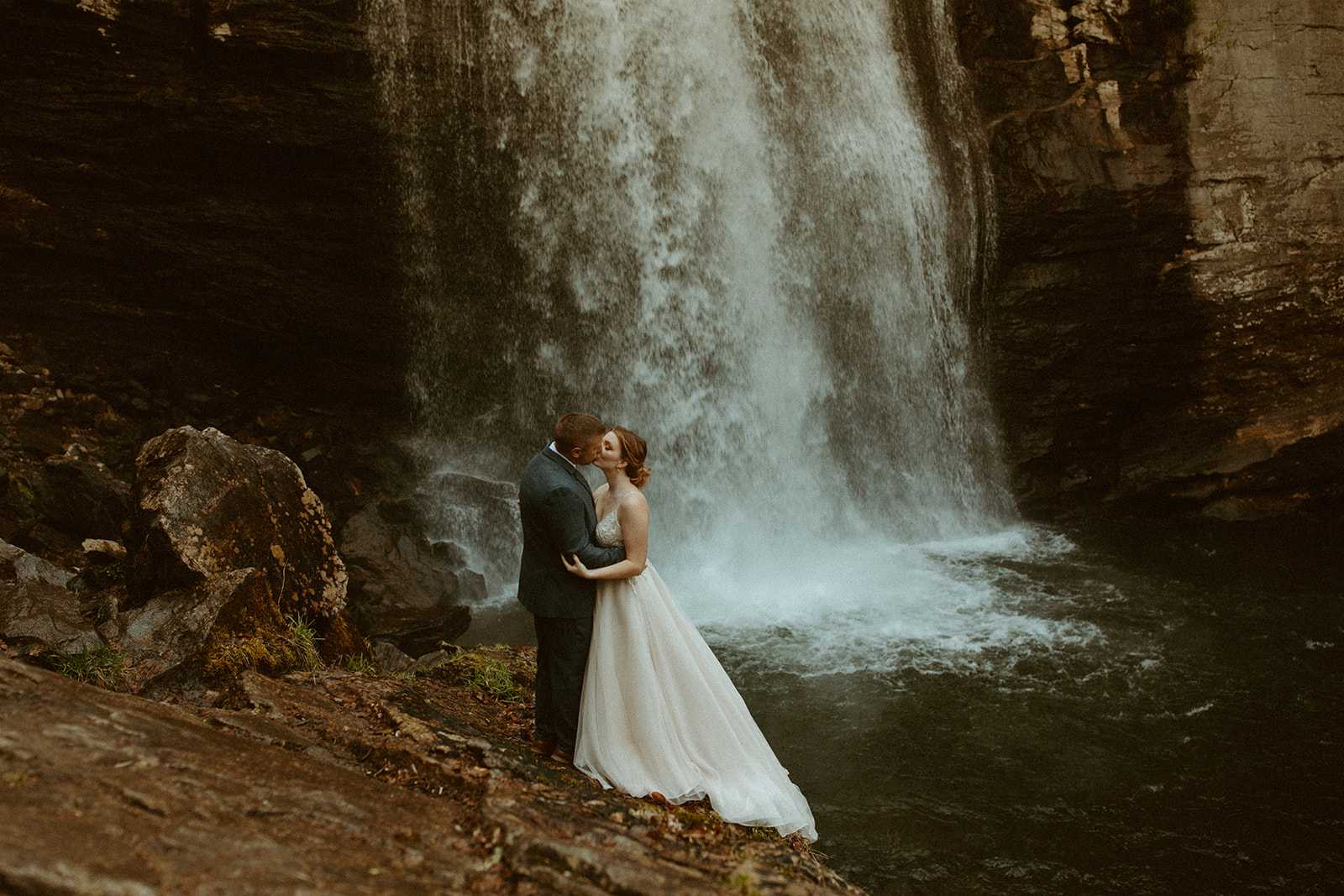 A couple who just got married stands near a waterfall embracing each other in their wedding attire.