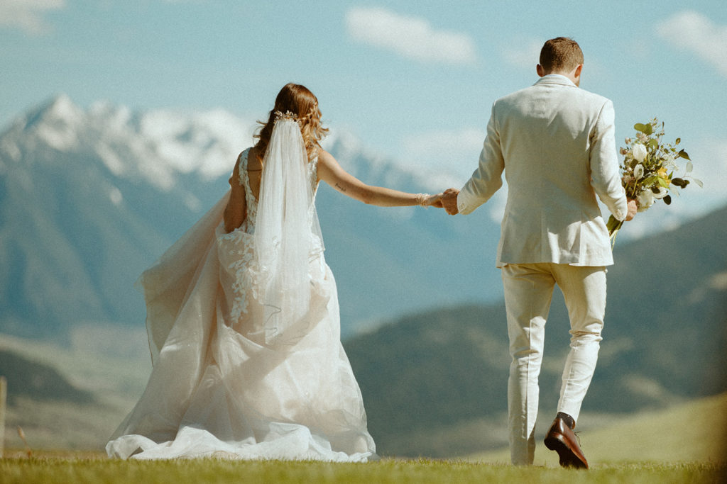 Bride and groom hold hands as they walk off in a meadow while the groom holds the bride's bouquet and mountains protrude in the background of their Montana elopement.