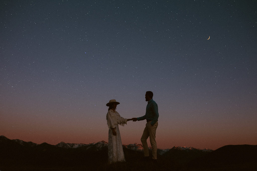 Bride and groom hold hands as their stare up at the stars while the sun has almost set during their elopement in Montana.