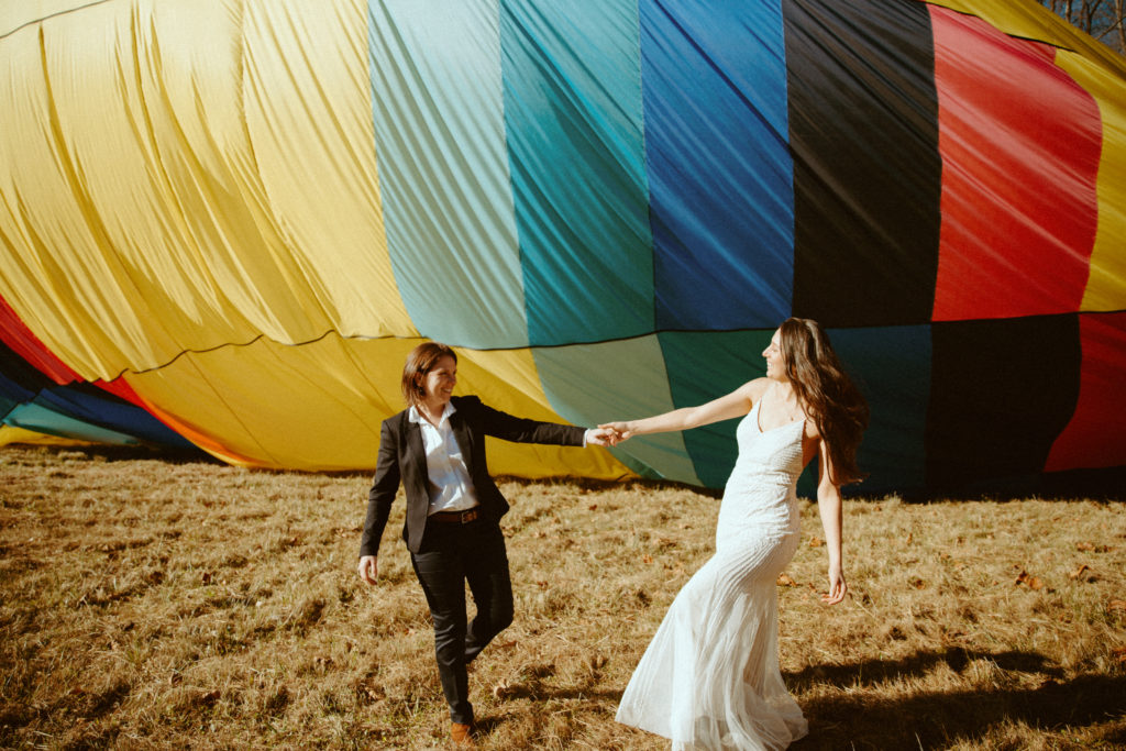 Two brides hold hands as they walk during their sunset elopement. They are walking in front of a deflating hot air balloon.