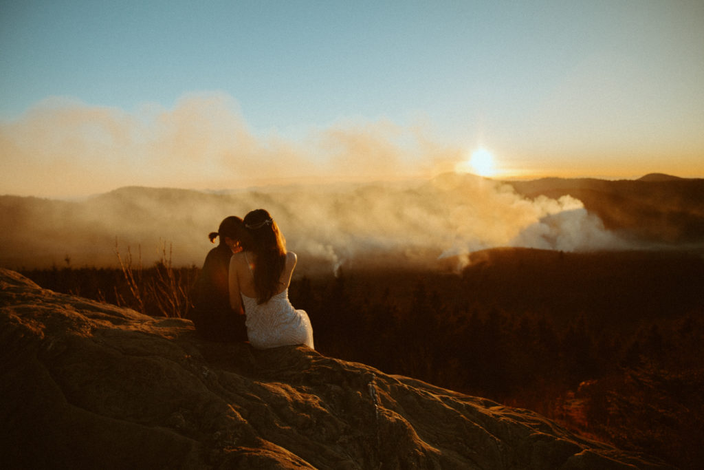 Two brides sit along side a ridge during a sunset elopement as they lean into one another. Fog begins to rise from the valley of trees below and the sun sets creating a glow.