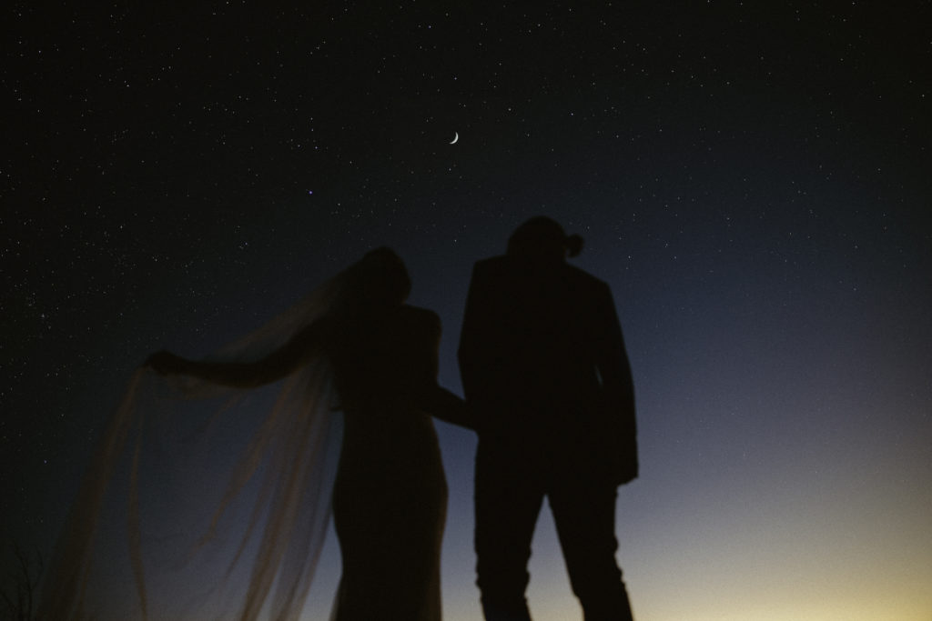Two brides in their wedding attire look up at the night sky with the stars shining above them.