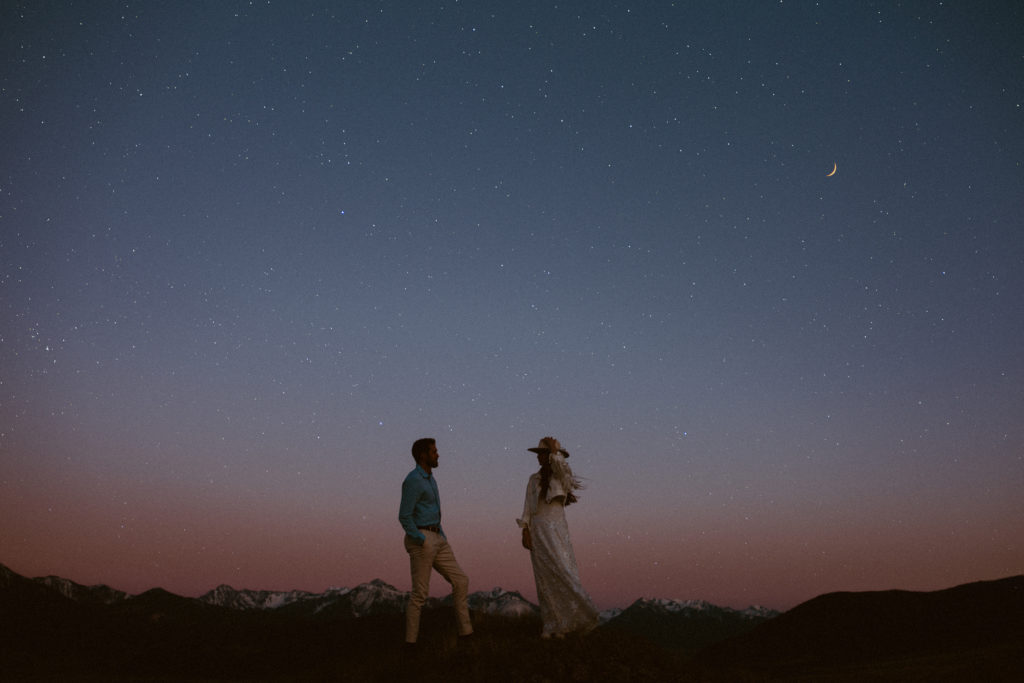 A couple, bride and groom, stand along a mountain ridge during the night with the stars shining above them.