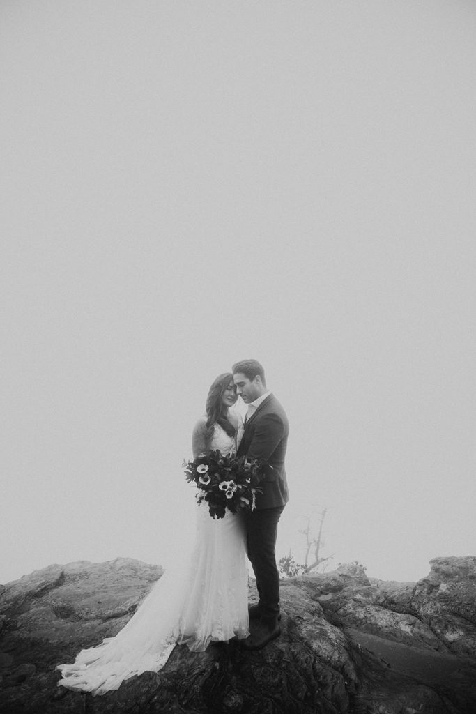 Black and white photo of a bride and groom embracing on a foggy mountain trail.