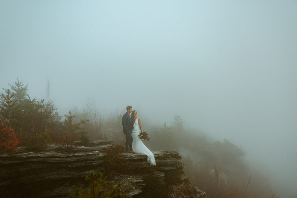 Bride and groom stare out over a foggy cliff.