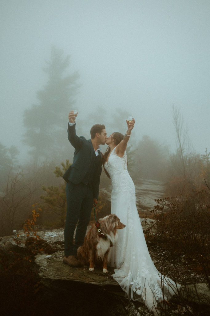 Bride and groom kiss while toasting with glasses on a mountain trail with their dog at their feet.