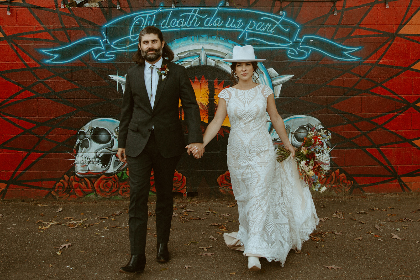 A bride and groom stand in front of a mural that says "Til death do us part."