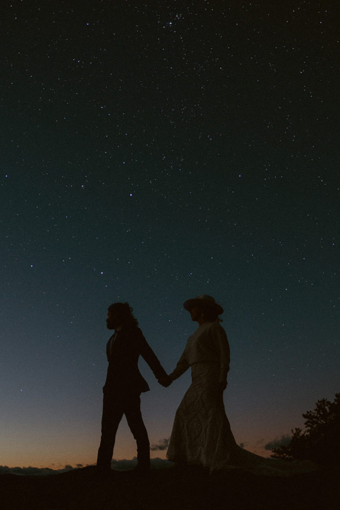 Bride and groom hold hands while walking and looking up at the sky filled with stars.