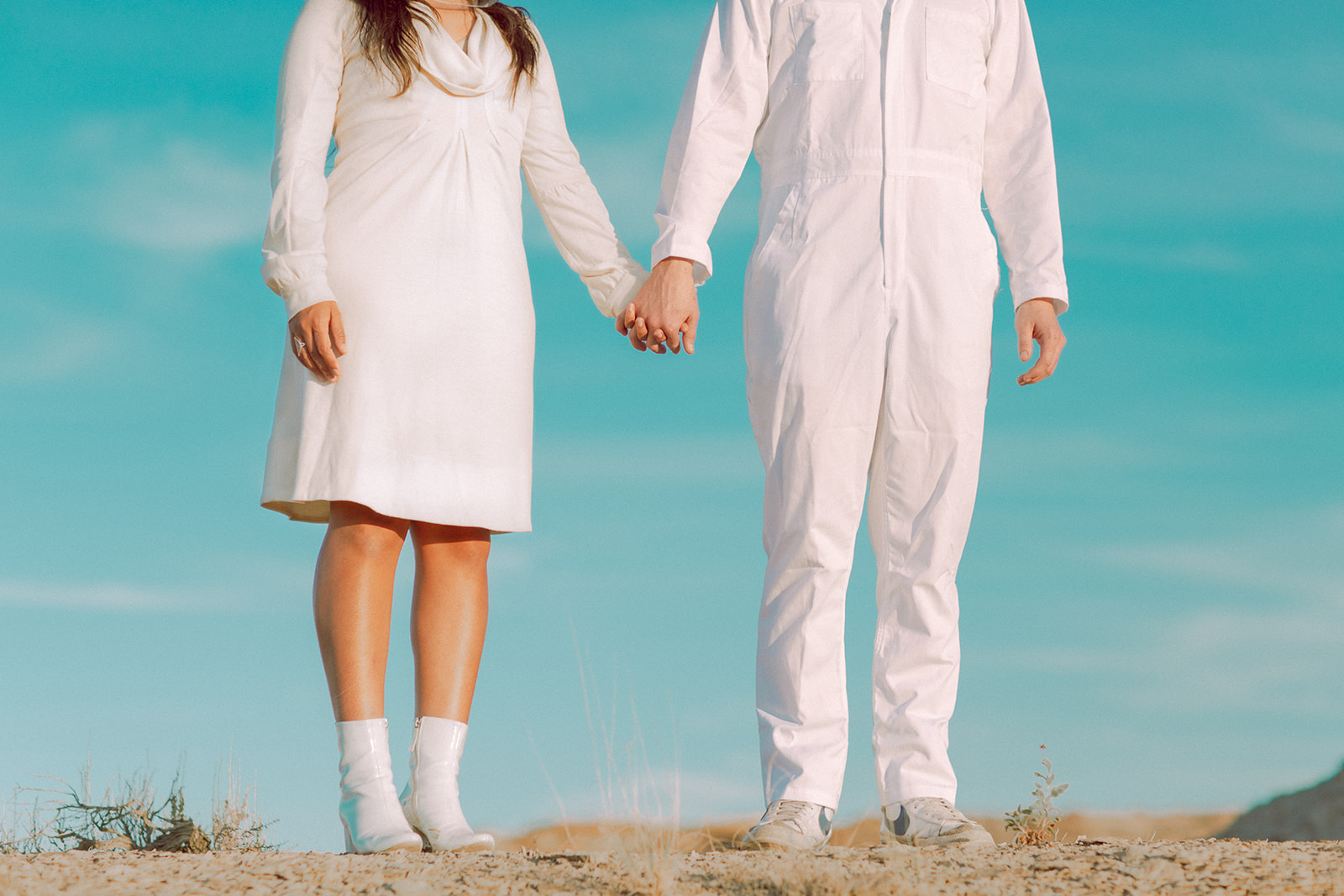 A man and woman hold hands while wearing all white outfits out in nature.